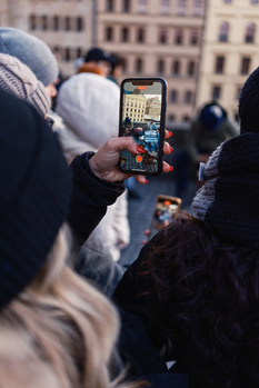 Person holding smartphone taking photo of crowded urban plaza with historic architecture in background.