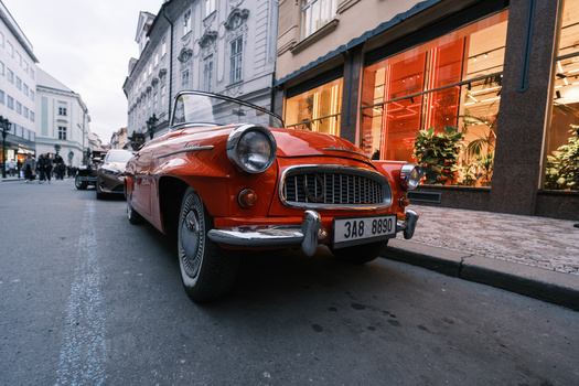 Red vintage convertible parked on a European city street with historic buildings and storefronts.