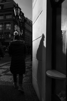 Person in dark coat walking away down an urban street with buildings and a storefront visible.