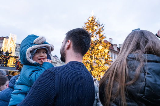 Man holding toddler in blue coat viewing illuminated Christmas tree at outdoor holiday market.