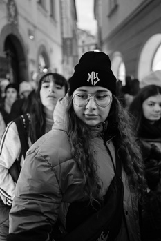 Woman with long dark hair and glasses wearing a beanie in a crowded urban street, looking directly at camera.