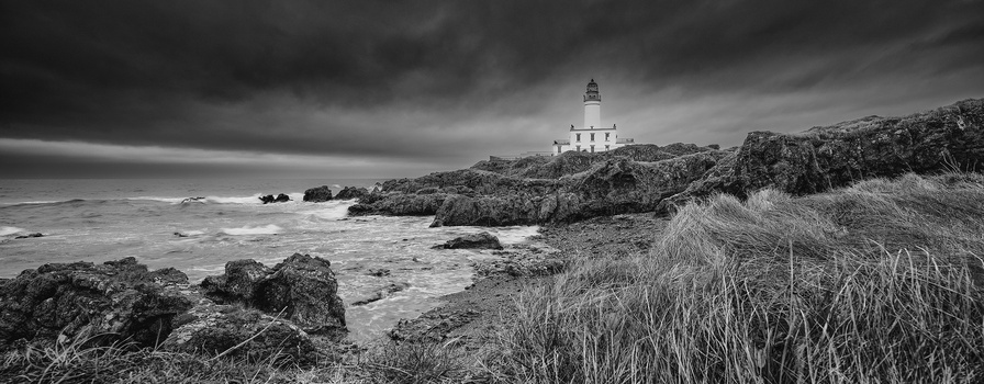 Lighthouse on rocky coastal headland with rough seas and dramatic storm clouds overhead.