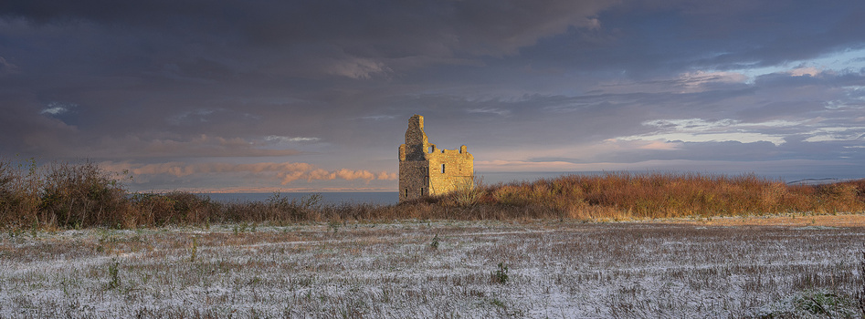 Ruined stone tower illuminated by golden sunset light in an expansive snow-covered field with bare trees.