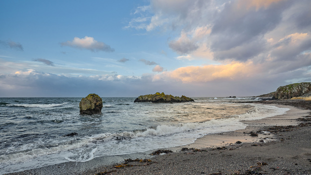 Rocky coastline with scattered sea stacks and breaking waves under a partly cloudy sky at golden hour.