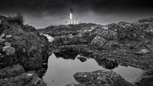 Lighthouse standing on rocky coastline with tidal pools reflecting dramatic storm clouds in black and white.