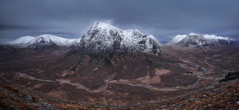 Snow-capped mountain peaks rise dramatically from a barren, winding valley floor under overcast skies.