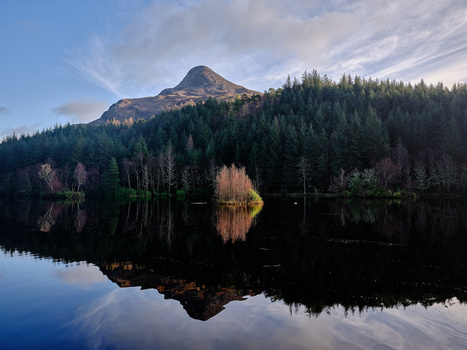 Mountain reflected in still lake surrounded by evergreen forest under partly cloudy sky.