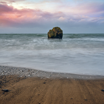 Rocky sea stack surrounded by foamy waves at twilight with soft pink and blue clouds overhead.