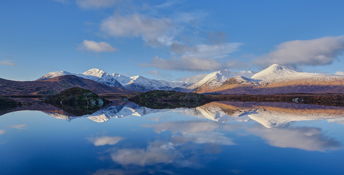Snow-capped mountains reflected in still water with golden moorland in foreground.