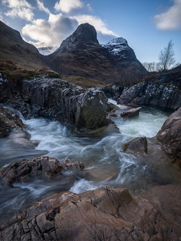 Mountain stream with flowing water over dark rocks, snow-capped peak rising in misty background.