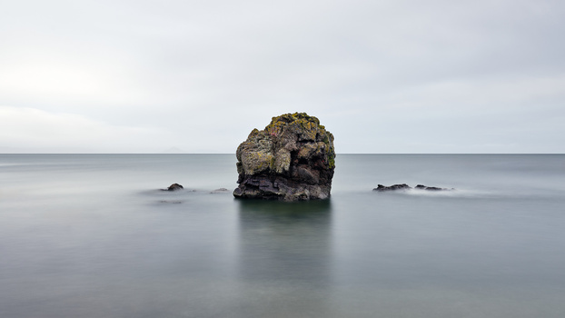 Solitary rock formation rising from calm shallow seawater with smaller rocks nearby.