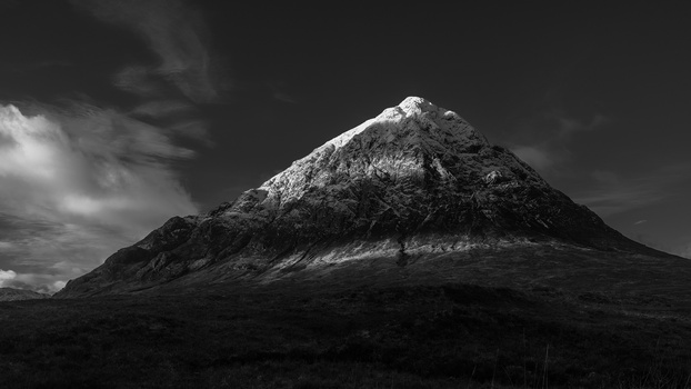 Snow-capped conical mountain rising dramatically from dark terrain under stormy sky in black and white.