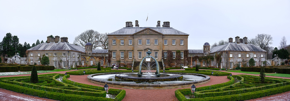 Stately brick mansion with symmetrical wings flanking a central fountain in formal garden setting.