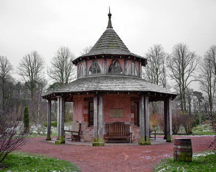 Red brick gazebo with conical roof and columns in a park setting surrounded by bare trees.