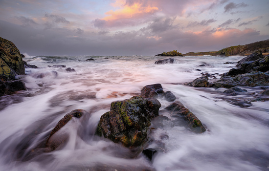 Rocky coastline at sunset with silky water motion blurring around moss-covered boulders.
