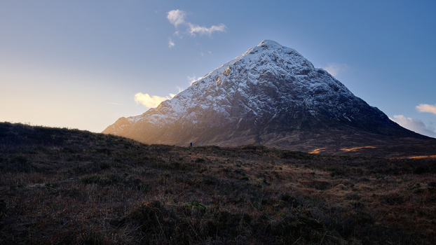 Snow-capped pyramid mountain rising from barren moorland under partly cloudy sky.