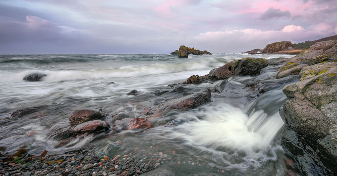 Rocky coastline with waves crashing over boulders under a dramatic cloudy sky at dusk.
