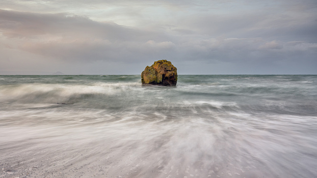 Solitary rock formation surrounded by crashing waves on a sandy beach under overcast sky.