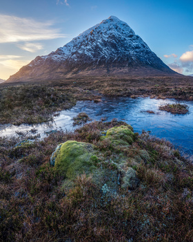 Snow-capped pyramidal mountain rising above moorland with pools of water reflecting blue sky.