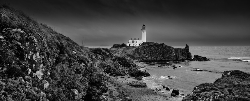 Lighthouse on rocky coastline under stormy sky in black and white.