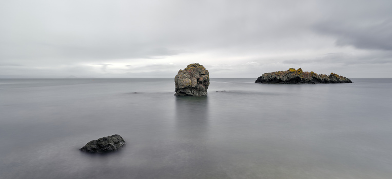 Rocky sea stacks rising from calm, misty ocean water under overcast sky.