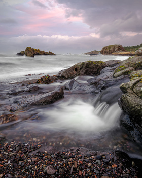 Rocky coastal landscape with long-exposure flowing water and dramatic cloudy sky at dusk.