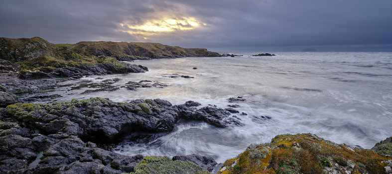 Rugged coastal landscape with moss-covered rocks in foreground, turbulent sea, and distant cliffs under dramatic storm clouds.