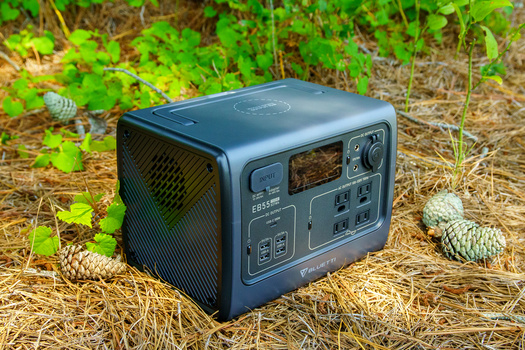 Portable power station with display screen and control panel sitting on ground surrounded by grass and plants.