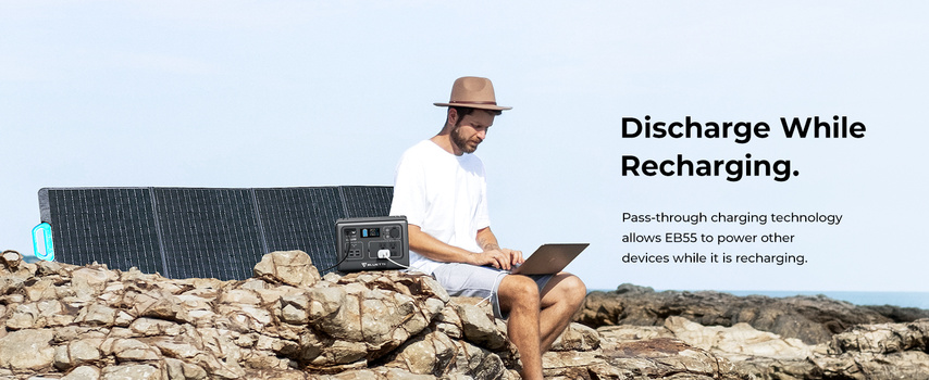 Man sitting on rocky terrain with solar panel and portable power station, using laptop outdoors.