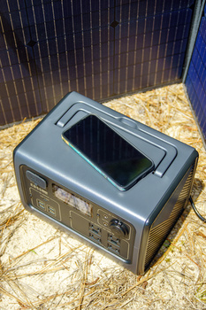 Portable power station with smartphone mounted on top, displayed on textured ground surface.