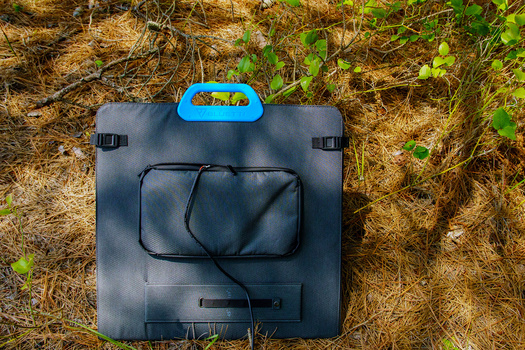 Solar panel charger with blue handle lying on dry grass and moss-covered ground.