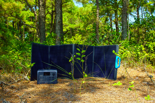Portable solar panels and power station set up on forest floor against dark corrugated metal backdrop.