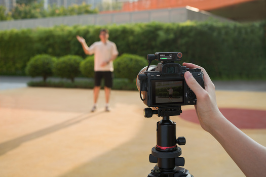 Camera operator filming a person standing in an outdoor courtyard with green hedges and architecture in the background.