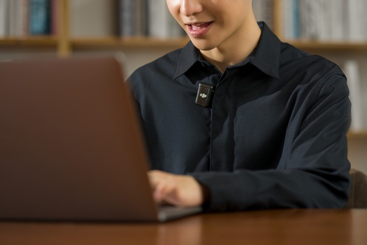 Person in black shirt using laptop at wooden desk with shelving in background.