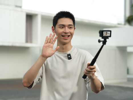 Young man waving at camera while holding a selfie stick with smartphone mounted on it.