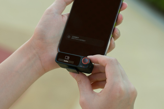 Hands holding a smartphone with an external lens adapter attachment mounted on the bottom.