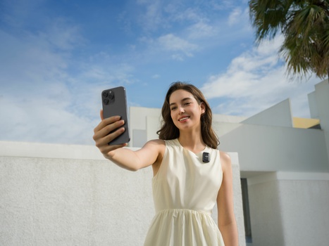 Woman in cream sleeveless dress taking a selfie with smartphone against modern white building.