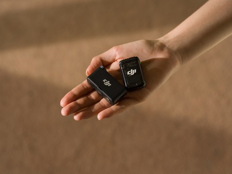 Hand holding two small black wireless microphone transmitters against a sandy background.