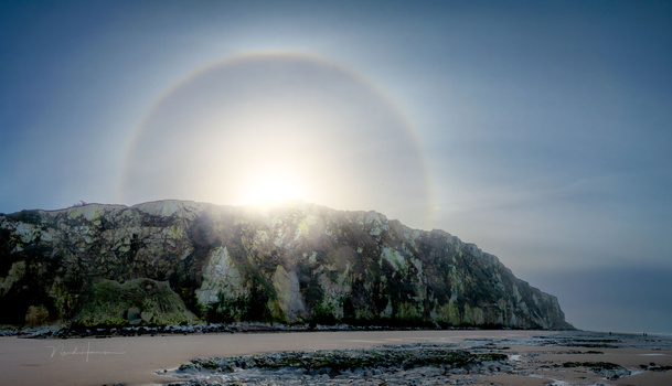 Rocky coastal cliff with bright sun creating a halo effect in blue sky above sandy beach.