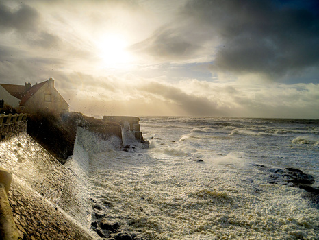 Coastal house and stone structure on cliff overlooking rough sea under dramatic golden-hour sky.