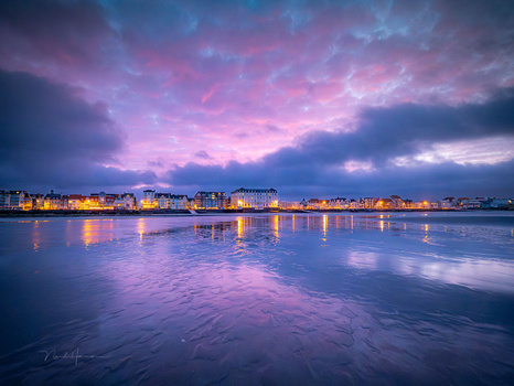 Frozen beach at twilight with pink and purple sky reflecting on icy sand and seaside town lights along the horizon.