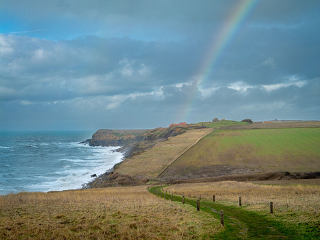 Coastal landscape with green fields, dramatic cliffs, and a rainbow stretching across stormy sky.