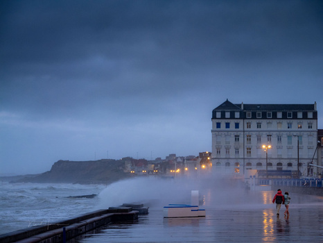 Coastal town at dusk with illuminated waterfront building and rough seas during blue hour.