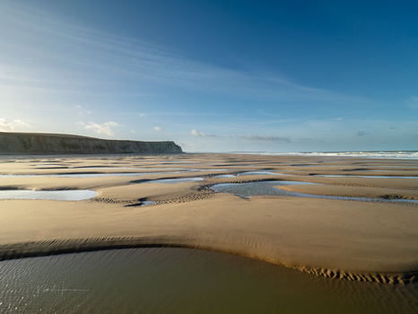 Expansive tidal beach with shallow water channels, rippled sand patterns, and distant cliffs under clear blue sky.