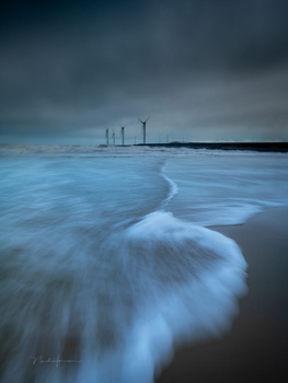 Wave washing onto a dark beach at dusk with industrial structures visible on the horizon under stormy skies.
