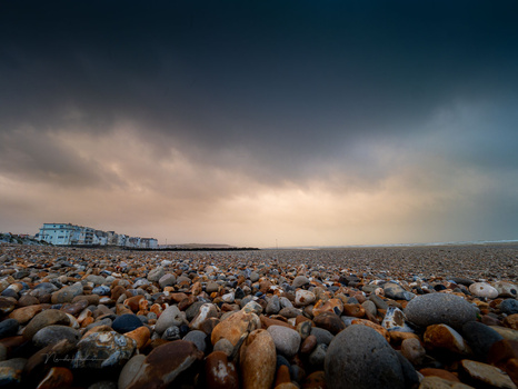 Pebble beach stretching toward a distant pier under dramatic cloudy sky.