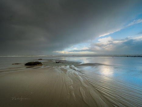 Sandy beach at twilight with rocky outcrops and calm shallow water reflecting dramatic cloudy sky.