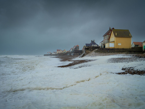 Yellow house on a rocky beach under stormy skies with rough waves and overcast conditions.