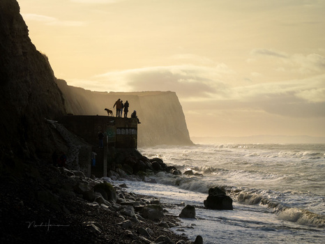 Silhouetted figures stand on a clifftop structure overlooking a dramatic coastline with crashing waves at golden hour.