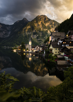 Alpine village nestled beside a lake with a church steeple, surrounded by steep forested mountains under dramatic cloudy sky.